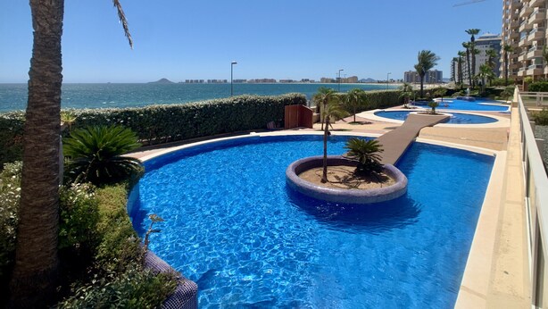 A swimming pool with a view of the sea, surrounded by palm trees and a building. - Bright and fully equipped in a front line development (La Manga del Mar Menor)