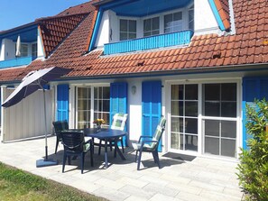 Una casa de dos pisos con techo de tejas, contraventanas azules y un patio con mesa y sillas.