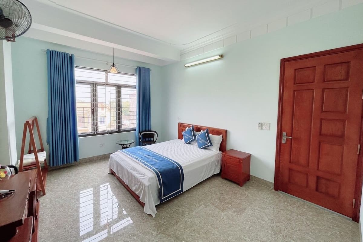 A bedroom with a bed, a desk, a chair, a window with curtains, a door, and a ceiling fan.