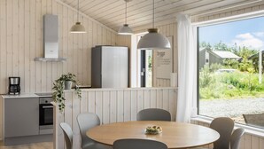 Eine moderne Küche mit Holzvertäfelung, einem Einbauofen und einem Kühlschrank. Ein Essbereich mit rundem Tisch und Stühlen lädt ein, und ein großes Fenster bietet Blick auf Grünflächen und ein Gebäude.