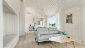 Ein modernes Wohnzimmer mit einem grauen Sofa, einem runden Holztisch und Blick ins Grüne durch große Fenster.