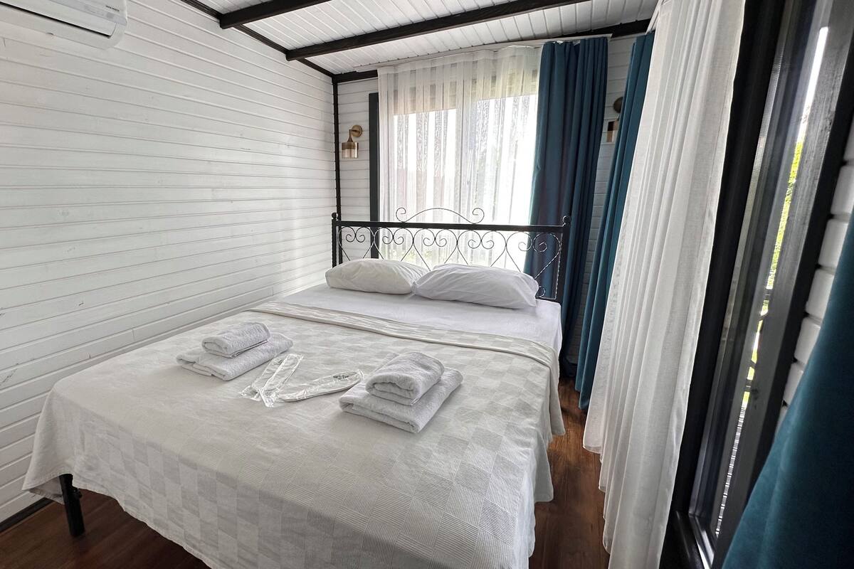A single bed with white linens and rolled towels, a window with sheer curtains, and a metal headboard.