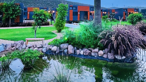 A modern building with a large water feature in the foreground, surrounded by greenery and a clear sky. - Flu Alaçatı Tiny House Otel (Çesme)