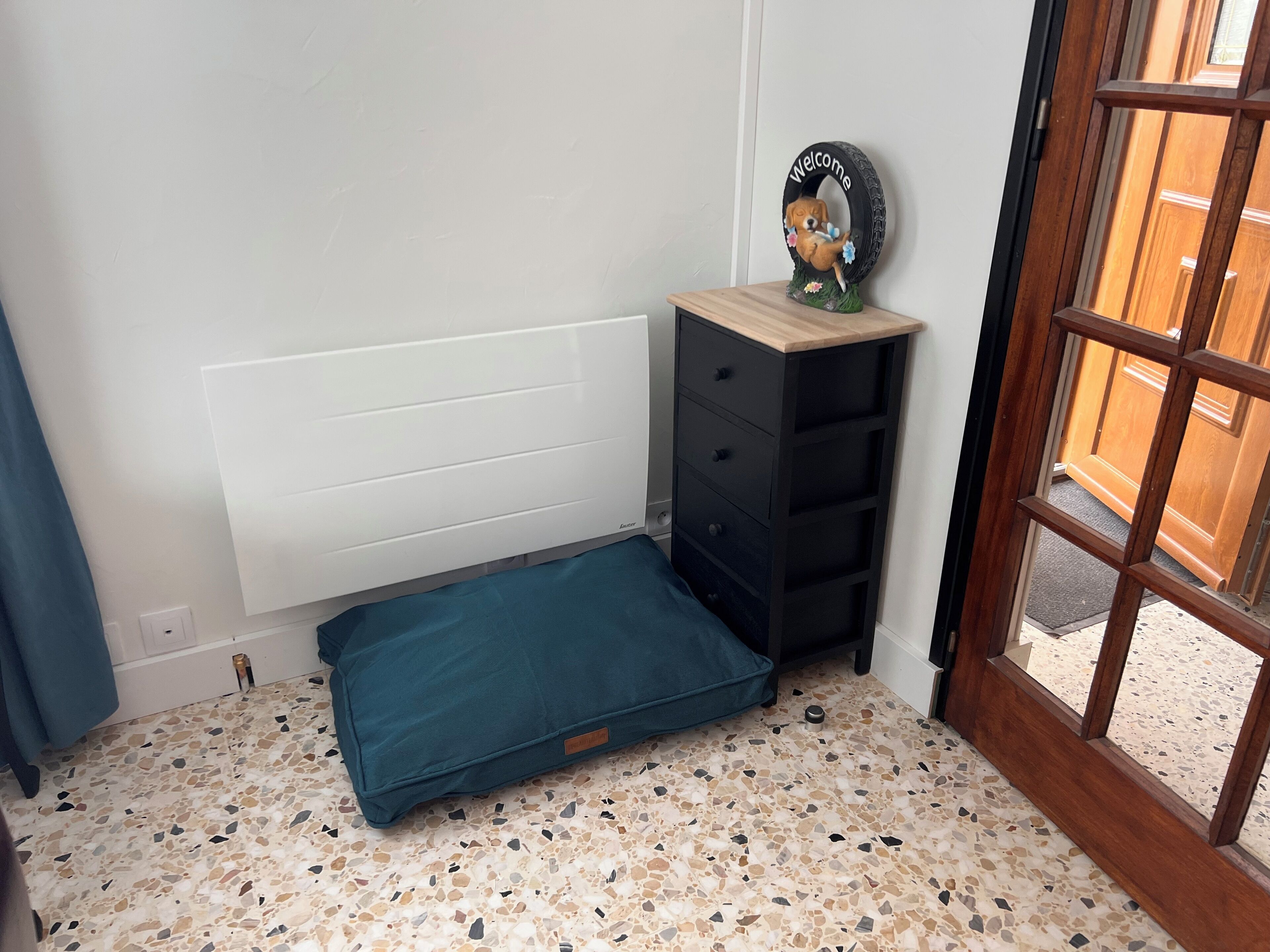 Een kamer met een bed, een klein zwart dressoir, een deurmat met decoratie van een hond, en een deur die naar buiten leidt.