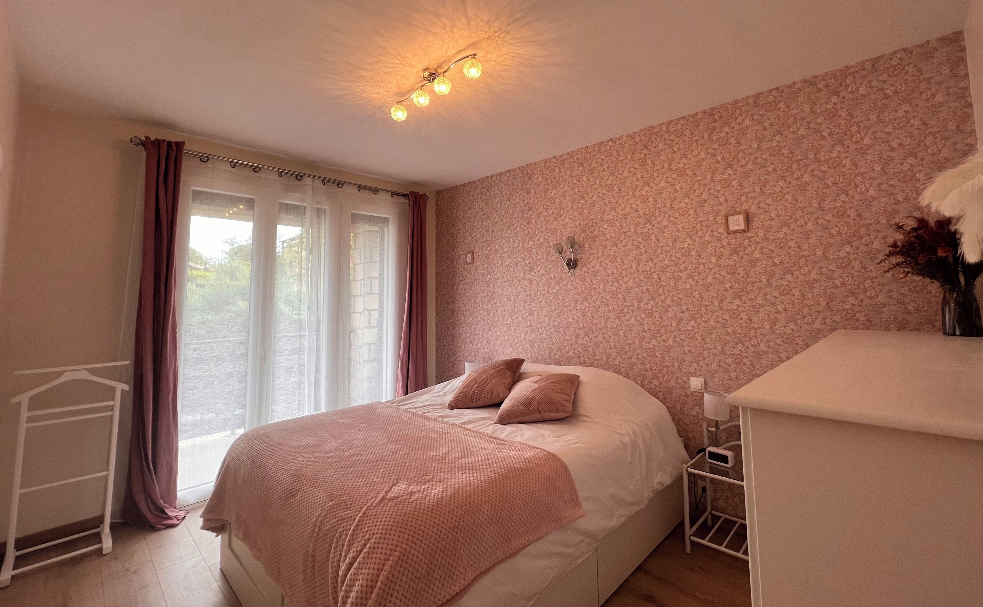 Een slaapkamer met een bed, nachtkastje, lamp, raam met gordijnen en een bloemen behang.