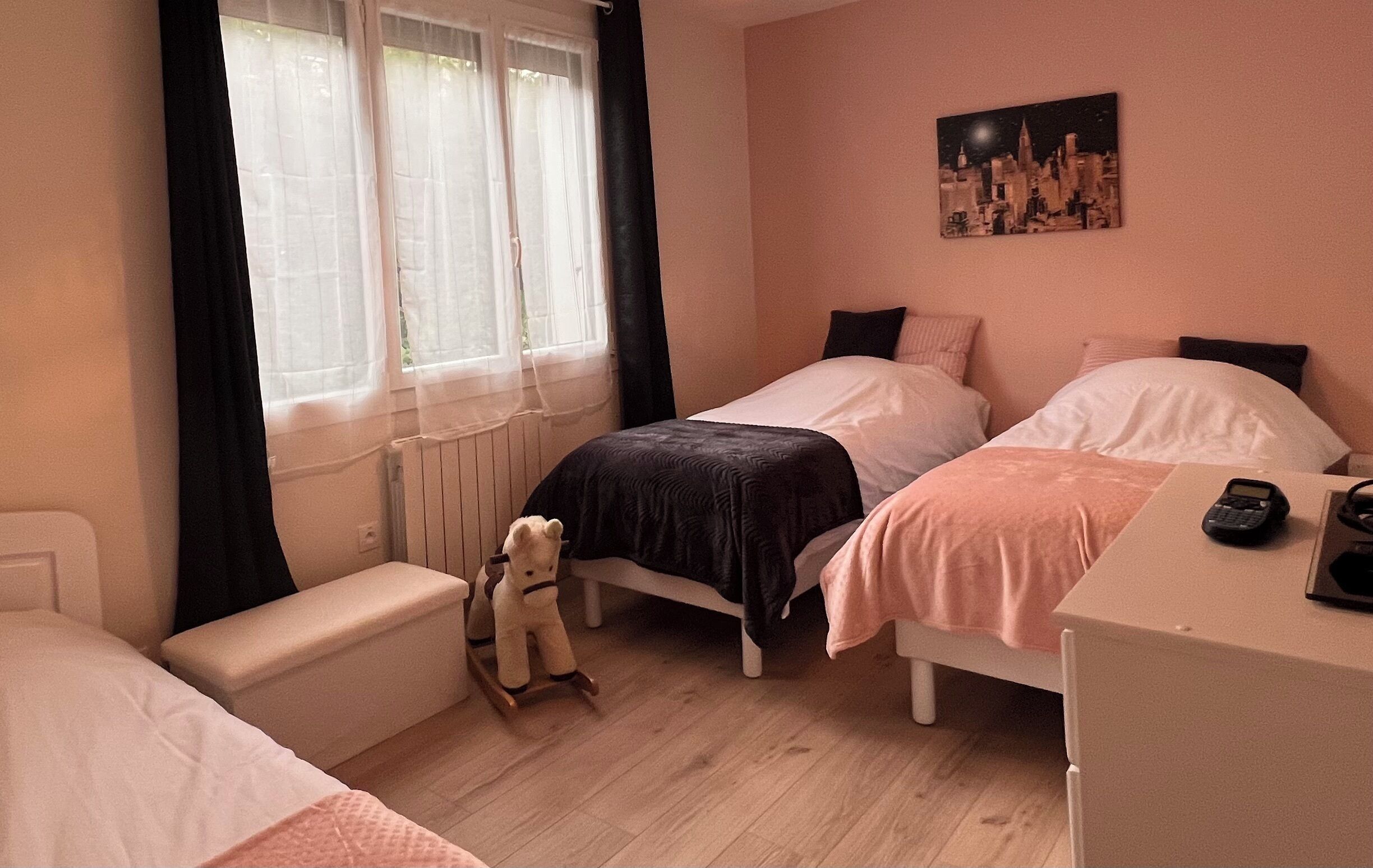 Ein Schlafzimmer mit zwei Betten, einem Fenster mit Vorhängen, einem Heizkörper, einem Teddybären, einem Schreibtisch mit Telefon und einem Stadtbild an der Wand.