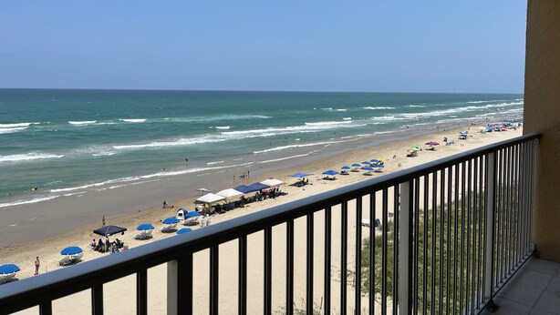 Une vue sur la plage depuis un balcon doté d’une balustrade, avec les vagues de l’océan et des parasols de plage.