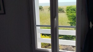 Een kamer met een raam dat uitkijkt op een strand en een grasveld. Er staat een witte stoel en een radiator bij het raam.