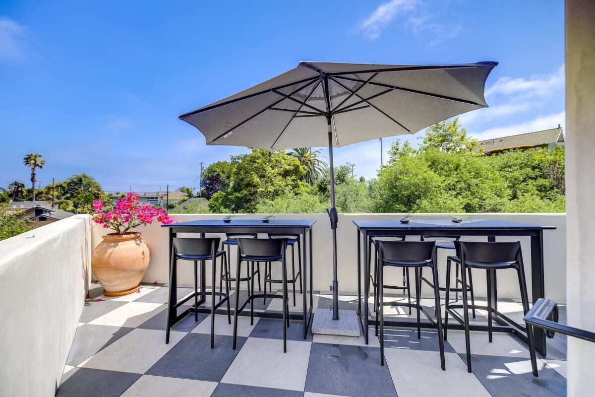 Eine Terrasse im Freien mit zwei Tischen, Stühlen, einem großen Sonnenschirm und einem Blumentopf mit rosa Blüten.