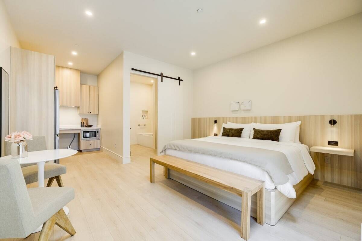 Una camera d'albergo moderna con un letto grande, una piccola scrivania, una panca e una kitchenette sullo sfondo.