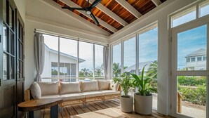 A sunlit room with a wooden bench, a sofa, and large windows offering a view of a house and palm trees.