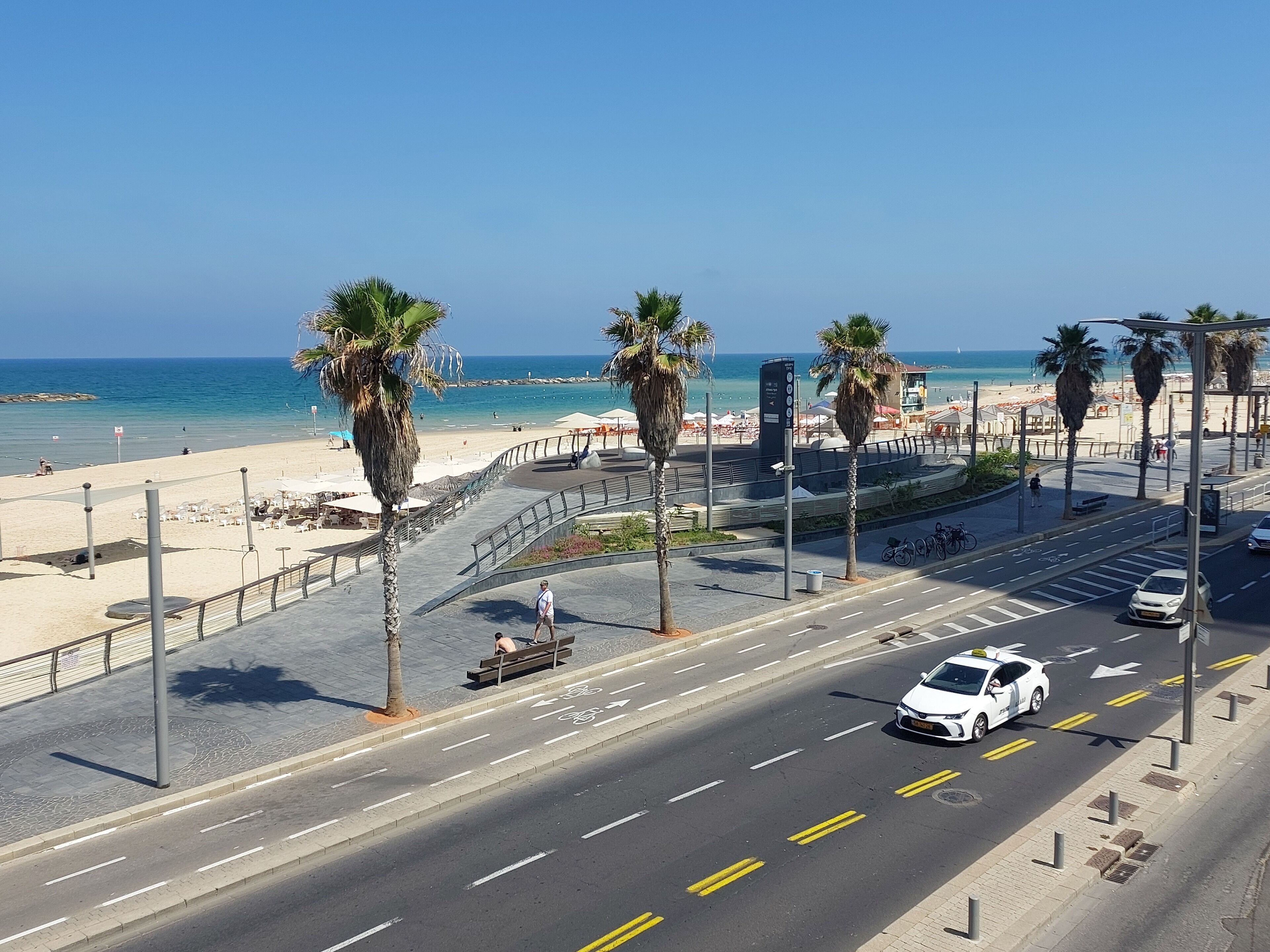 Una carretera costera con palmeras, una playa y un cielo azul despejado.