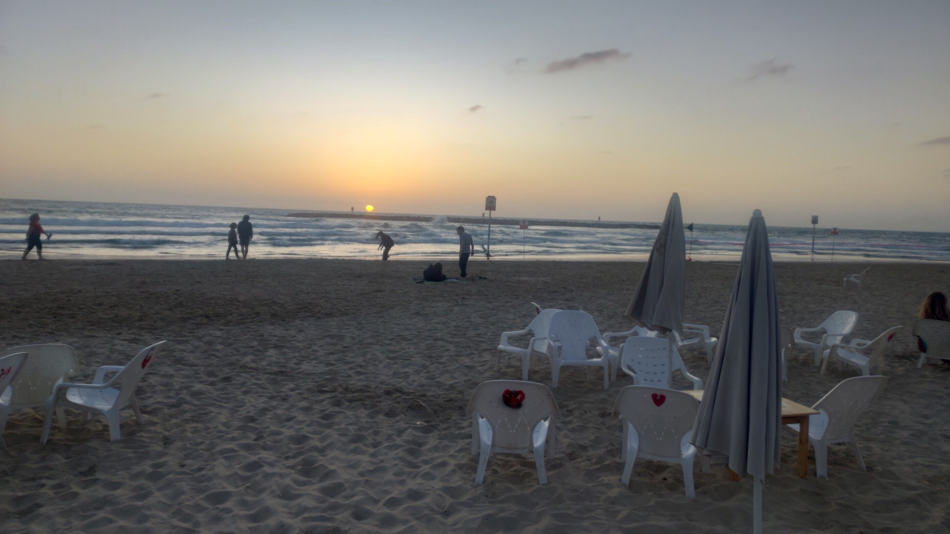 Una playa con sillas y sombrillas de plástico blanco, un atardecer en el fondo y gente caminando por la orilla.