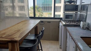 Een kleine keuken met een houten tafel en stoelen, een gasfornuis, een magnetron en een broodrooster. Een raam toont een gebouw met meerdere verdiepingen en balkons.