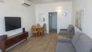 Een woonkamer met een flatscreen televisie, een houten eettafel met stoelen, een grijze bank en een schilderij aan de muur.