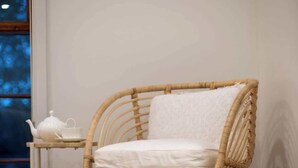 Une chaise en osier avec un coussin, une petite table avec une théière et une tasse, et un tapis à motifs posé sur un sol recouvert de moquette.
