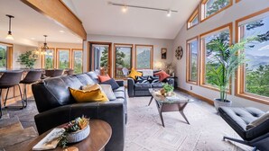 A spacious living room with a large sofa, a coffee table, and a dining area with chairs. Large windows offer a view of the outdoors.