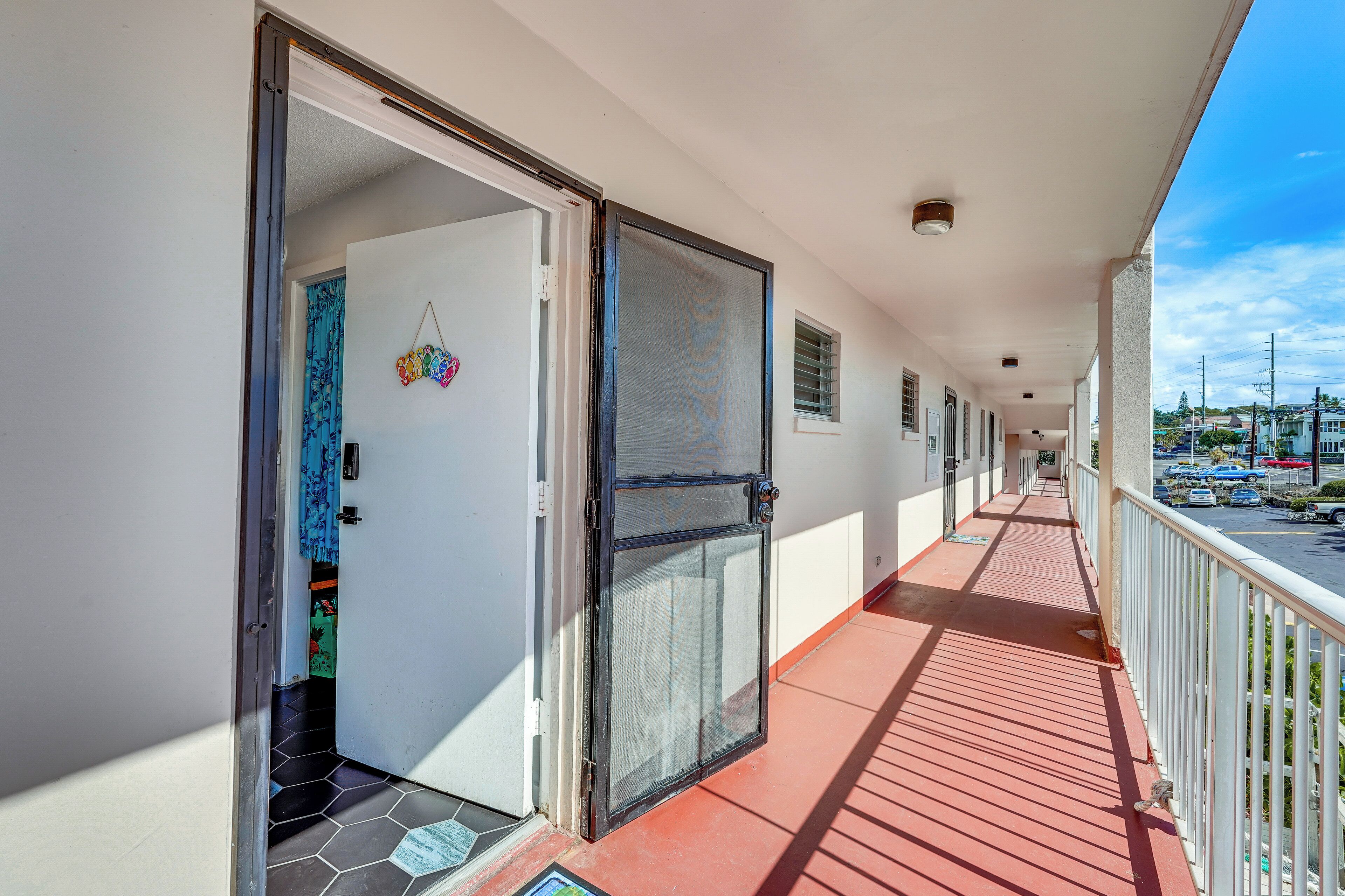 A covered balcony with a red floor, a white wall, and a door leading to a room.