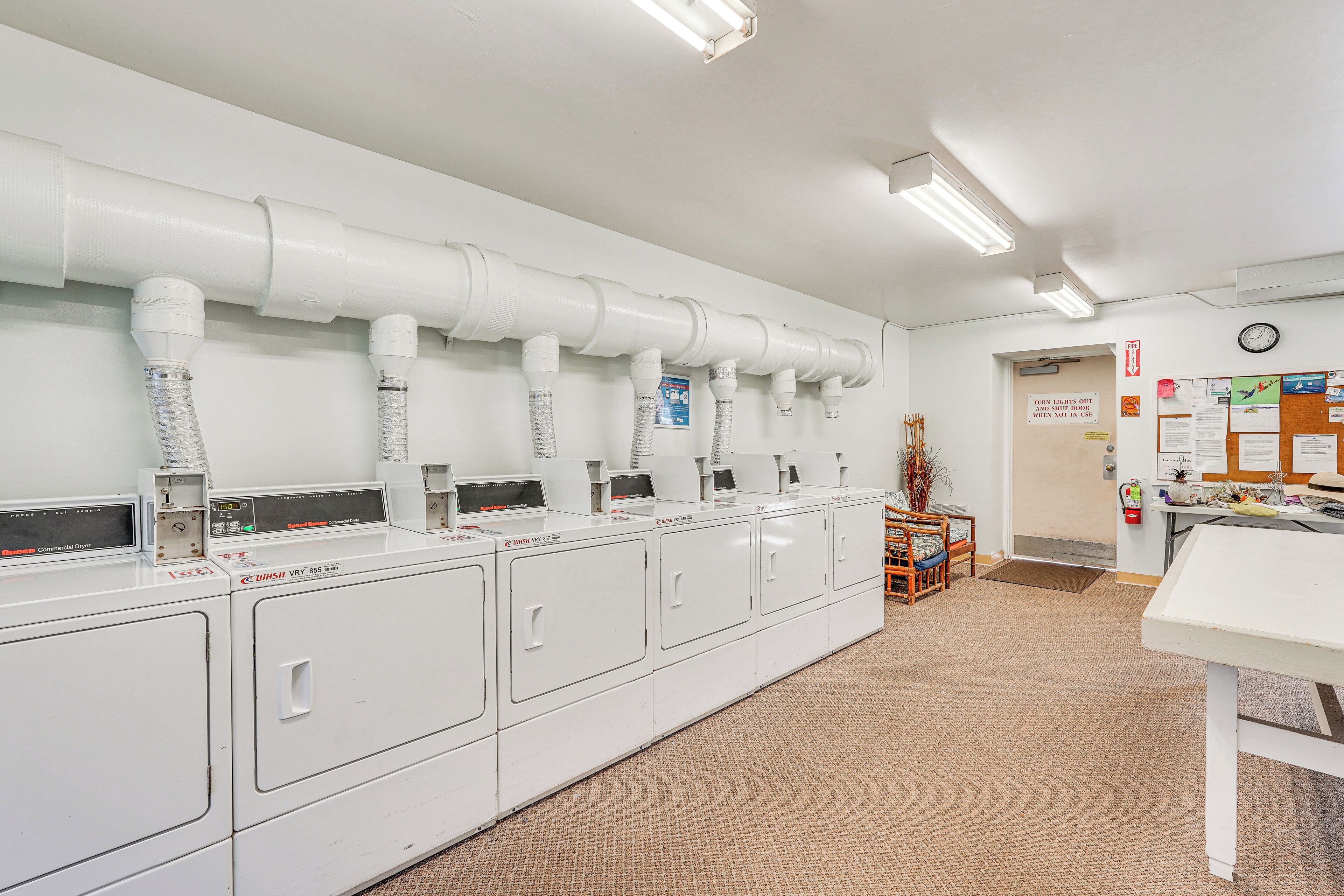 A laundry facility with multiple washing machines and dryers, a clock, and a bulletin board on the wall.