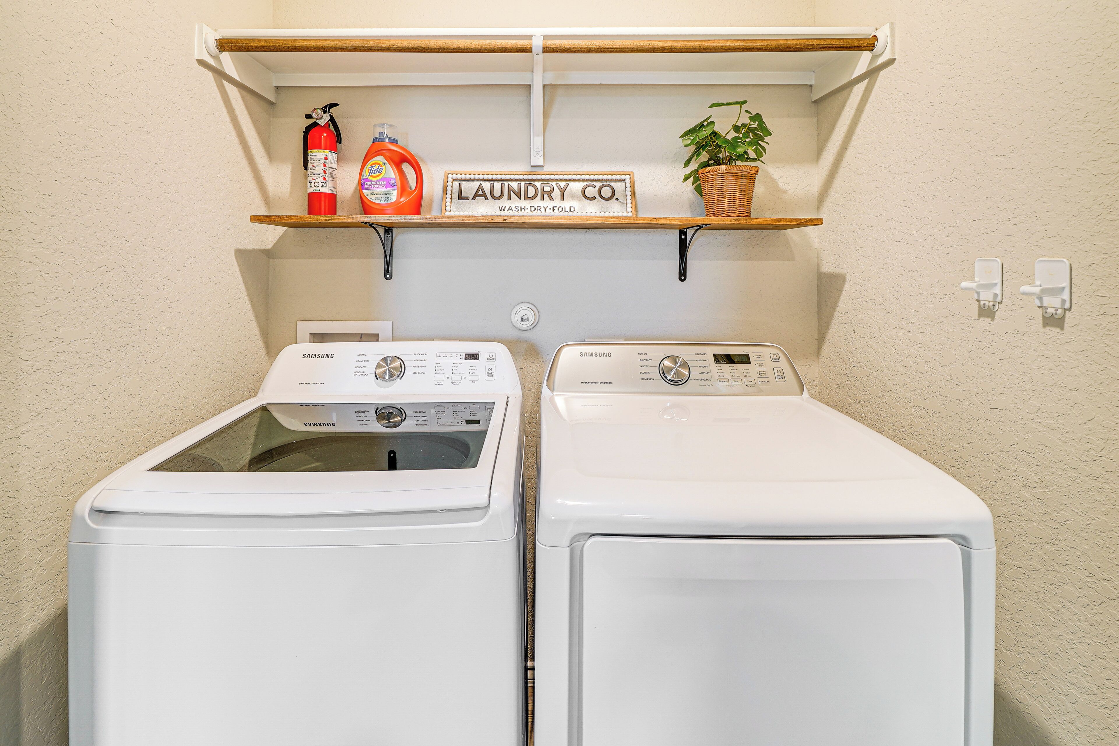 A laundry area with two front-loading washing machines, a shelf with cleaning supplies, a sign reading 'LAUNDRY CO.', and a potted plant.