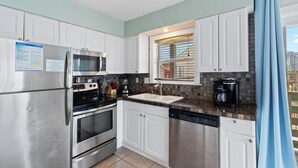 A modern kitchen with stainless steel appliances, white cabinetry, a built-in oven, and a window with blinds. - Gulf View 23 (Gulf Shores)
