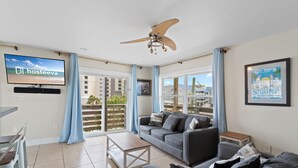 A modern living room with a sofa, coffee table, and a TV mounted on the wall. There is a ceiling fan, large windows with curtains, and a framed picture on the wall. - Gulf View 23 (Gulf Shores)