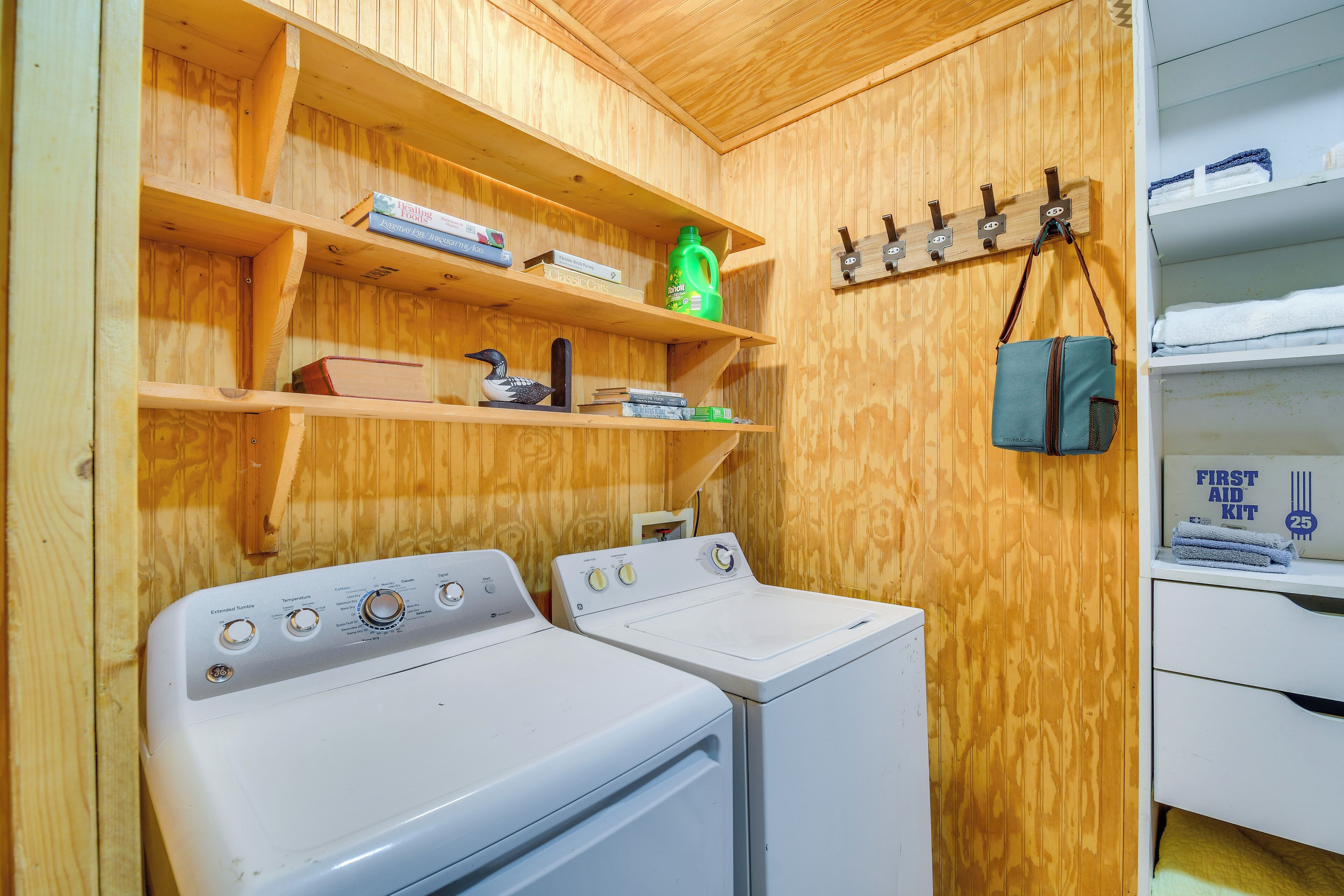 A laundry room with two washing machines, wooden shelves with books and a green bottle, and a hanging bag.