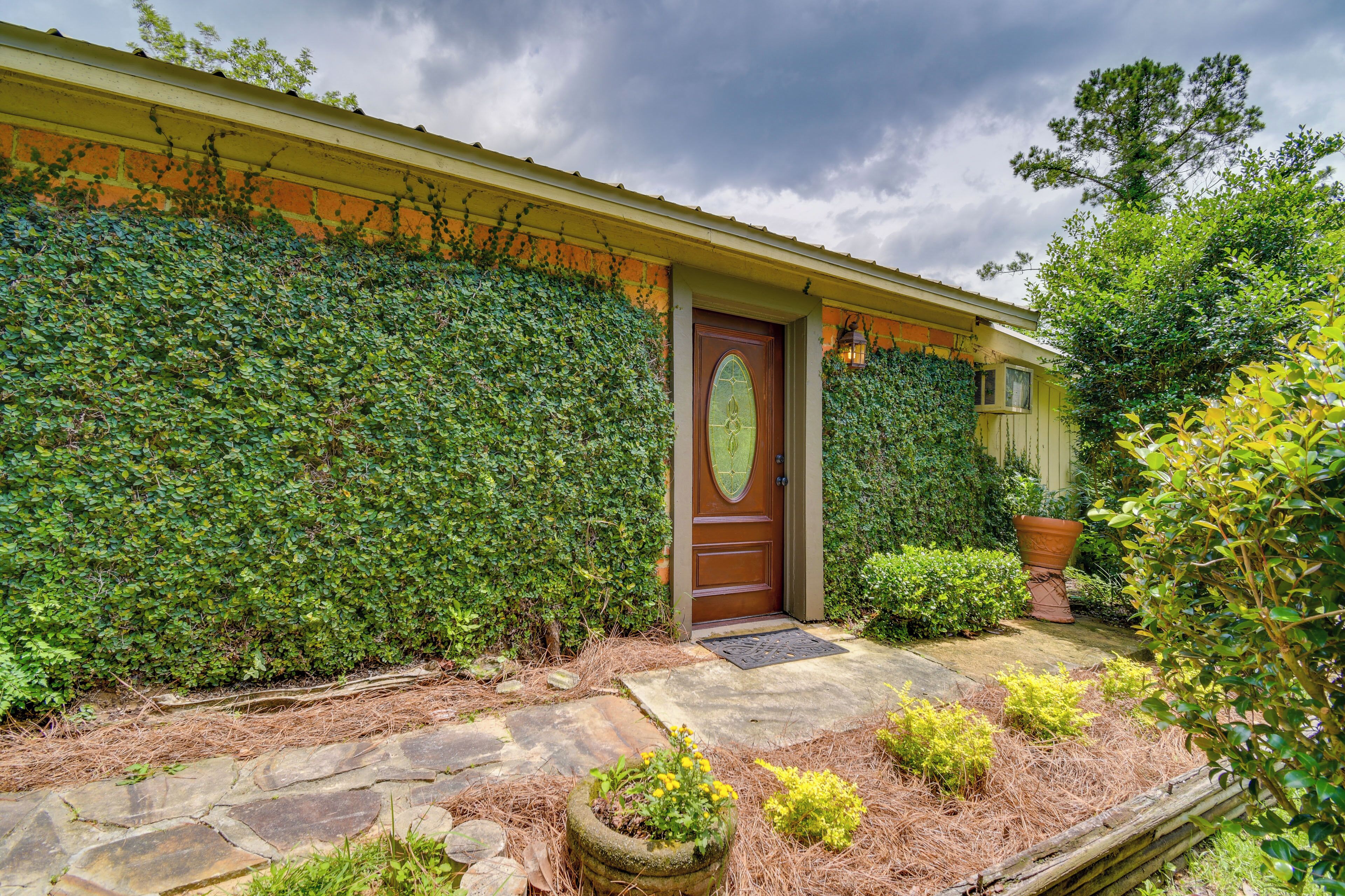 Une maison avec une porte en bois, un chemin de pierres et un jardin bien entretenu, agrémenté de diverses plantes et d’un grand bac à fleurs.
