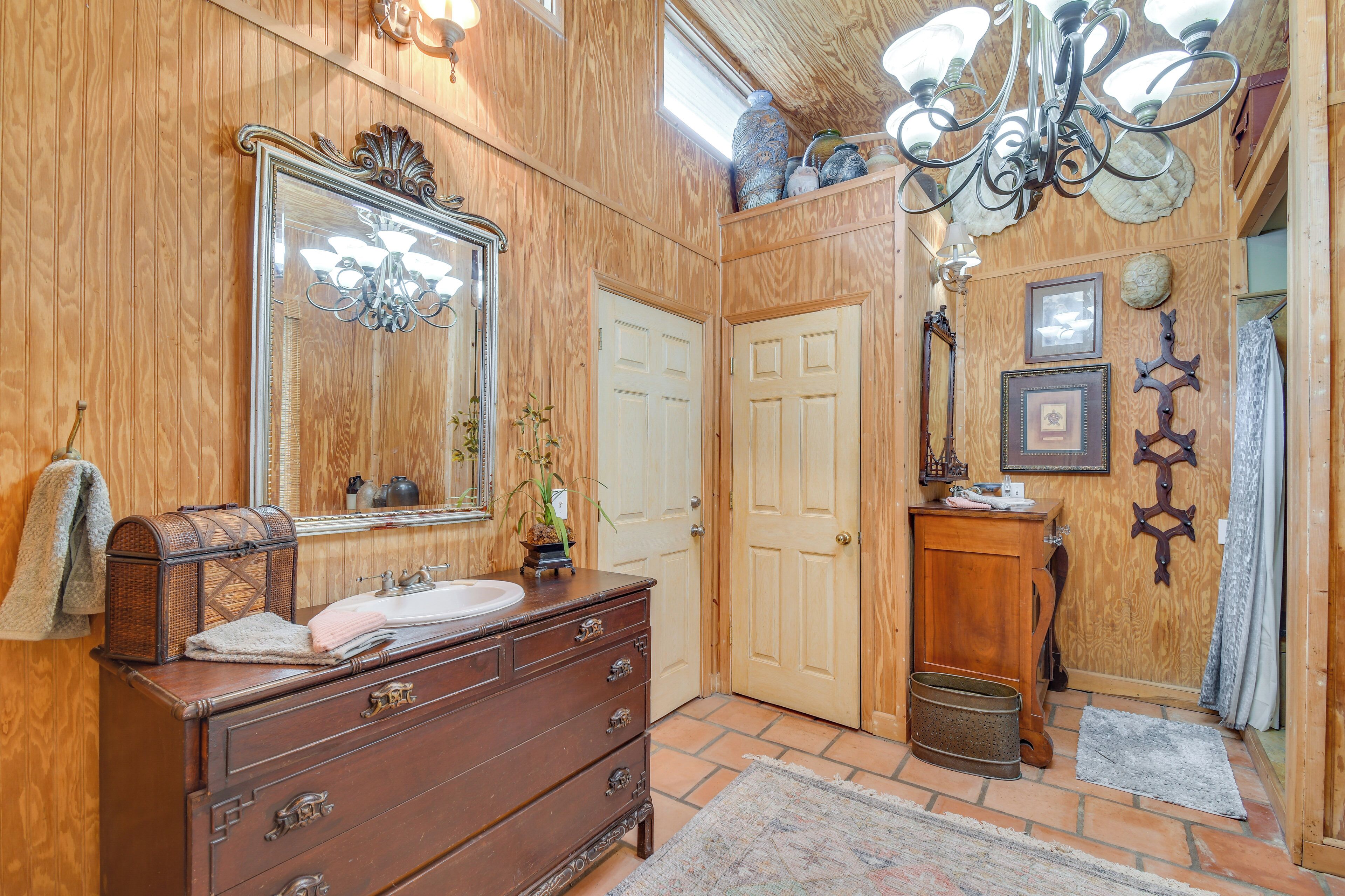 Une salle de bain aux murs lambrissés, dotée d’un grand miroir, d’une commode en bois, d’un lustre et d’une porte.