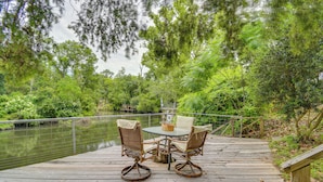 Una terrazza in legno con tavolo e sedie che si affaccia su uno specchio d'acqua, circondata da vegetazione lussureggiante.