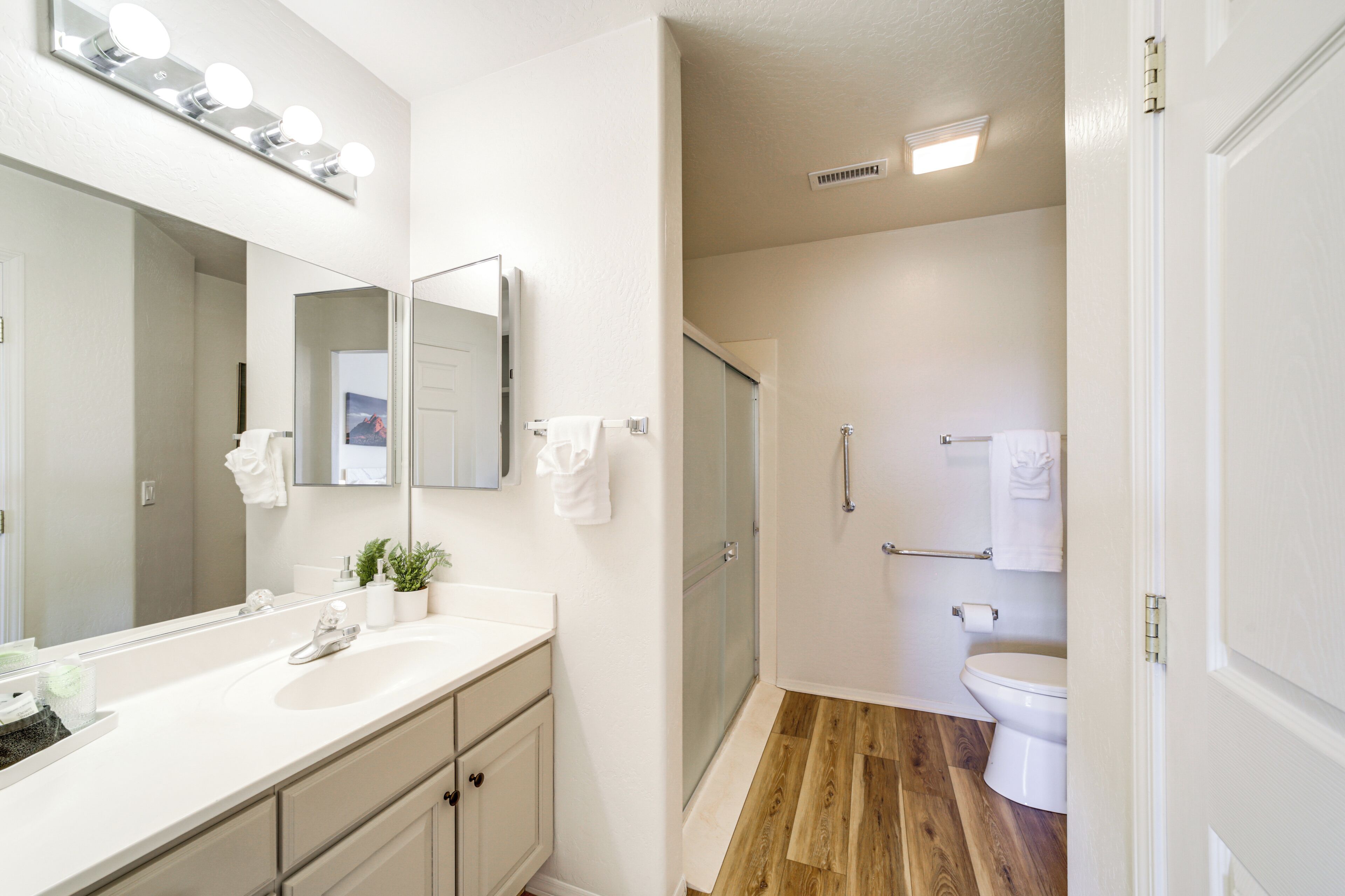 A bathroom with a white vanity, a large mirror with lights, a shower, and a toilet.