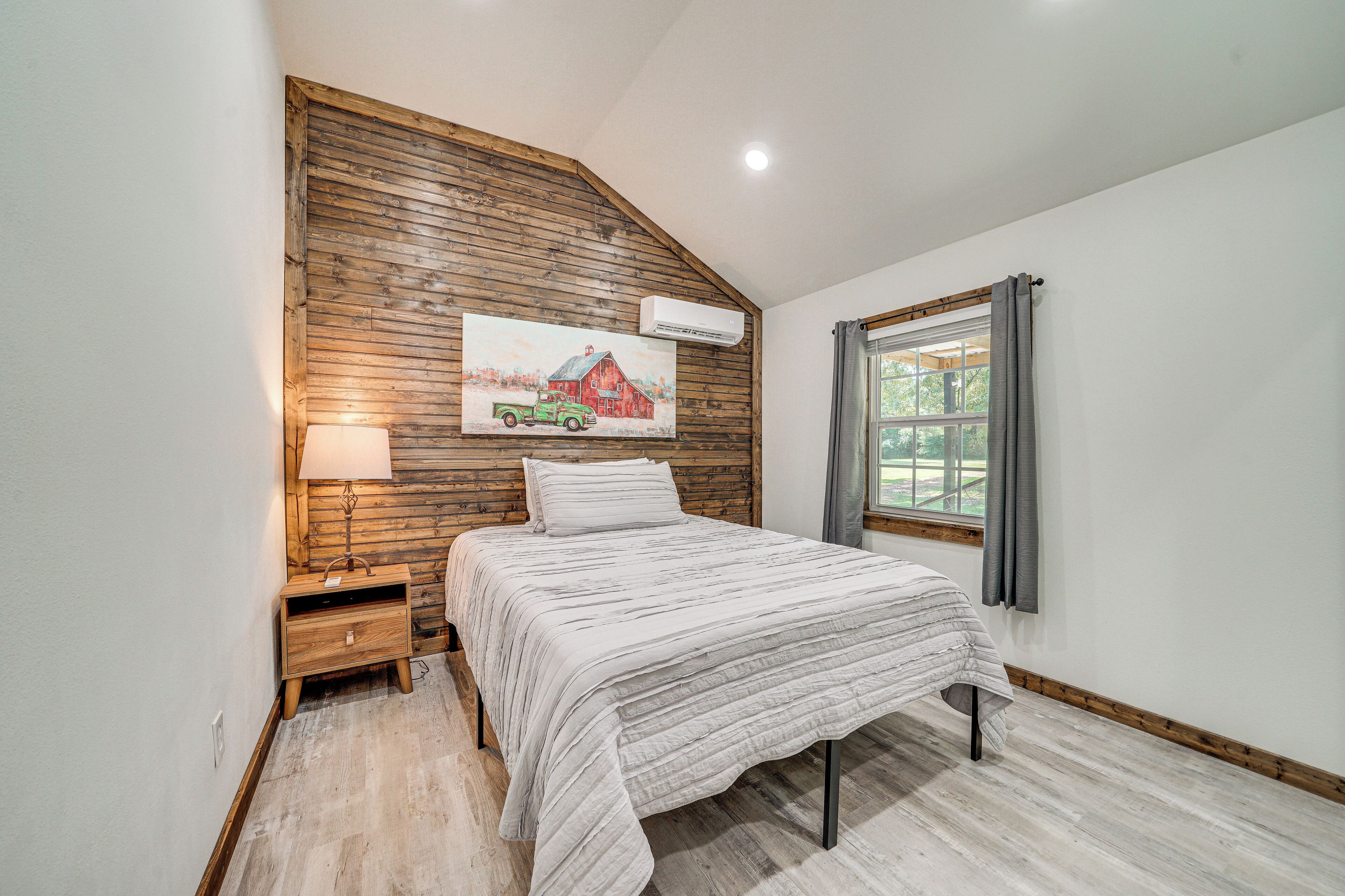 Un dormitorio con techo inclinado, cama con cubrecama blanco, mesita de noche con lámpara, panel de pared de madera y ventana con cortinas.