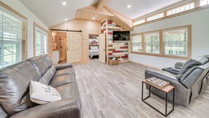 A spacious living area with a leather sofa, a recliner, and a small table. A wooden staircase leads to a loft bedroom with a bed and a TV. - Secluded Broken Bow Cabin w/ Fishing Pond & Views! (Broken Bow)
