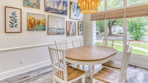 A dining area with a round wooden table, white chairs, and a large window with blinds.