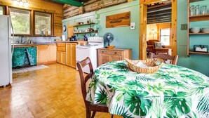 A rustic cabin interior with wooden walls, a dining table with a tropical leaf tablecloth, a kitchen with wooden cabinets, and a large window.