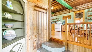 A wooden interior with a staircase leading to a dining area, shelves with kitchenware, and a kitchen in the background.