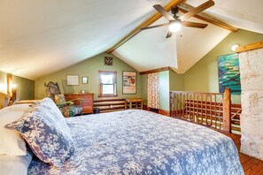 Une chambre avec un grand lit, une tête de lit en bois et un ventilateur de plafond. Il y a une fenêtre avec des rideaux et un tableau au mur.