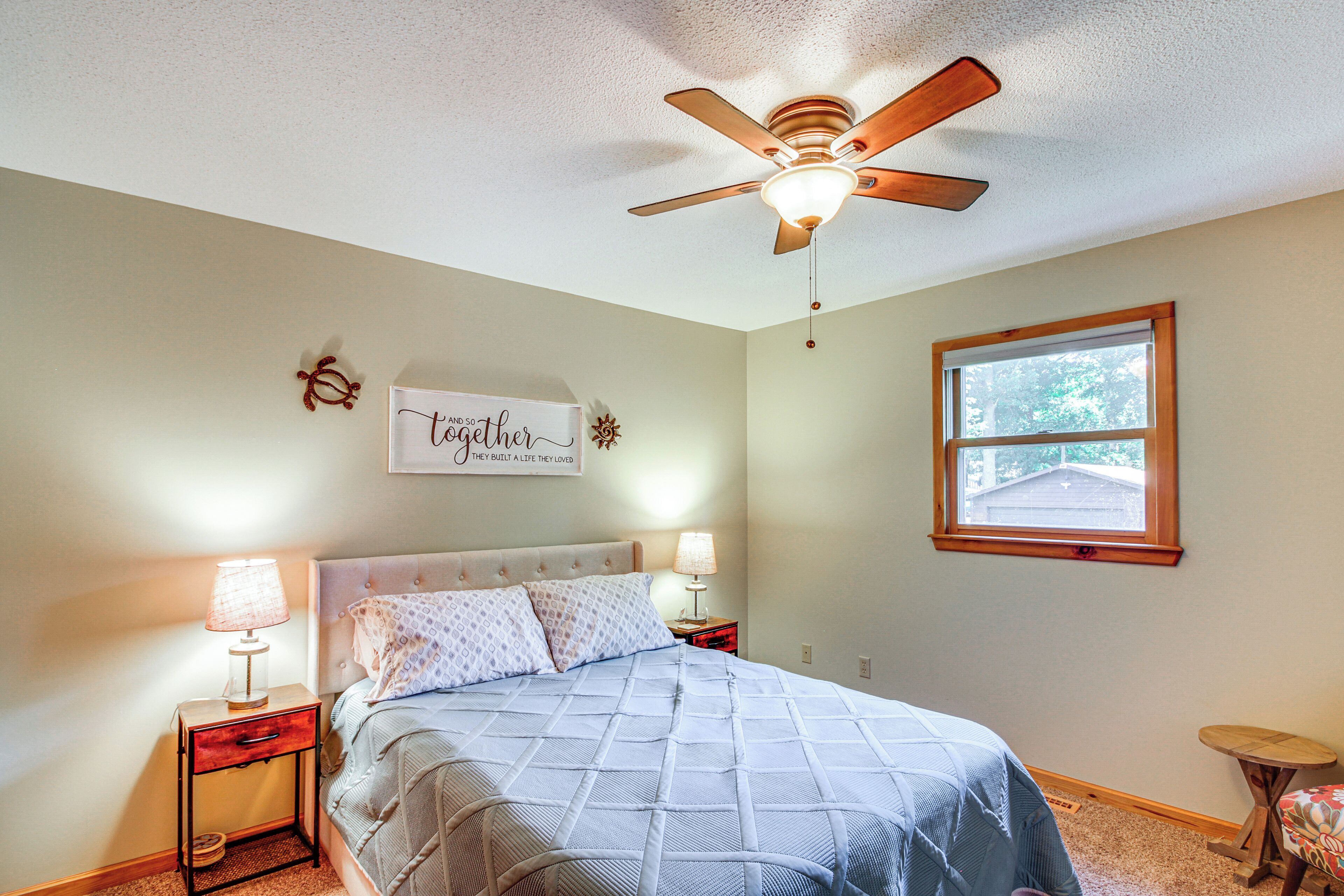 A bedroom with a bed, two bedside tables with lamps, a ceiling fan, a window, and a wall sign.