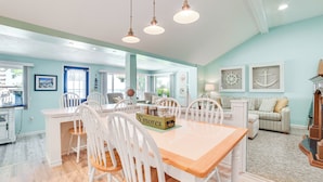 A spacious dining area with a wooden table and white chairs, a kitchen in the background, and a living area with a striped sofa and nautical decor. - Private Dock & Fire Pit: Houghton Lake Home! (Houghton Lake)