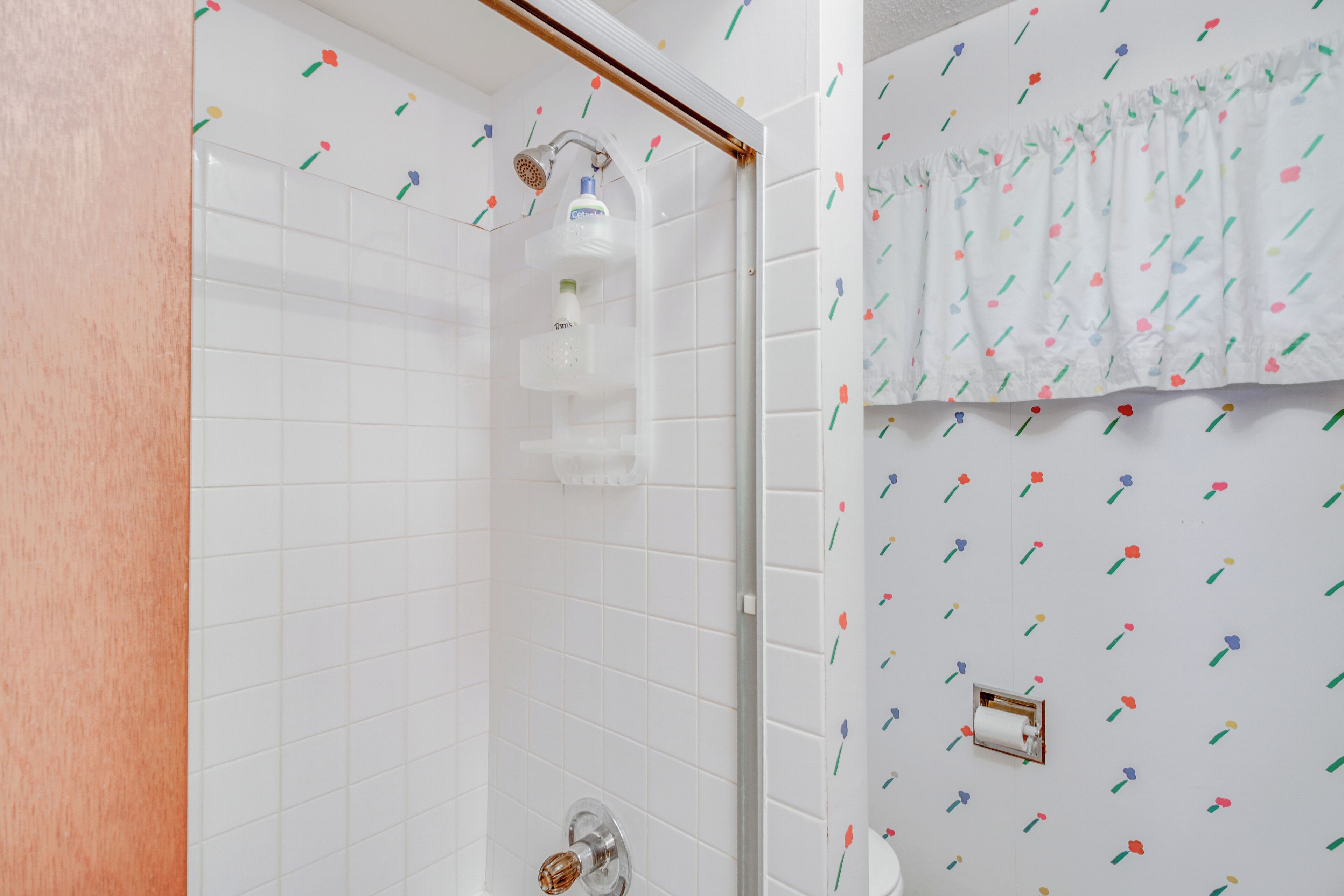 Un baño con paredes de azulejos blancos, ducha con puerta de cristal y un inodoro con cortina de estampado floral.