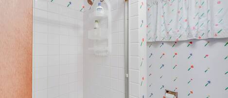 Un baño con paredes de azulejos blancos, ducha con puerta de cristal y un inodoro con cortina de estampado floral.