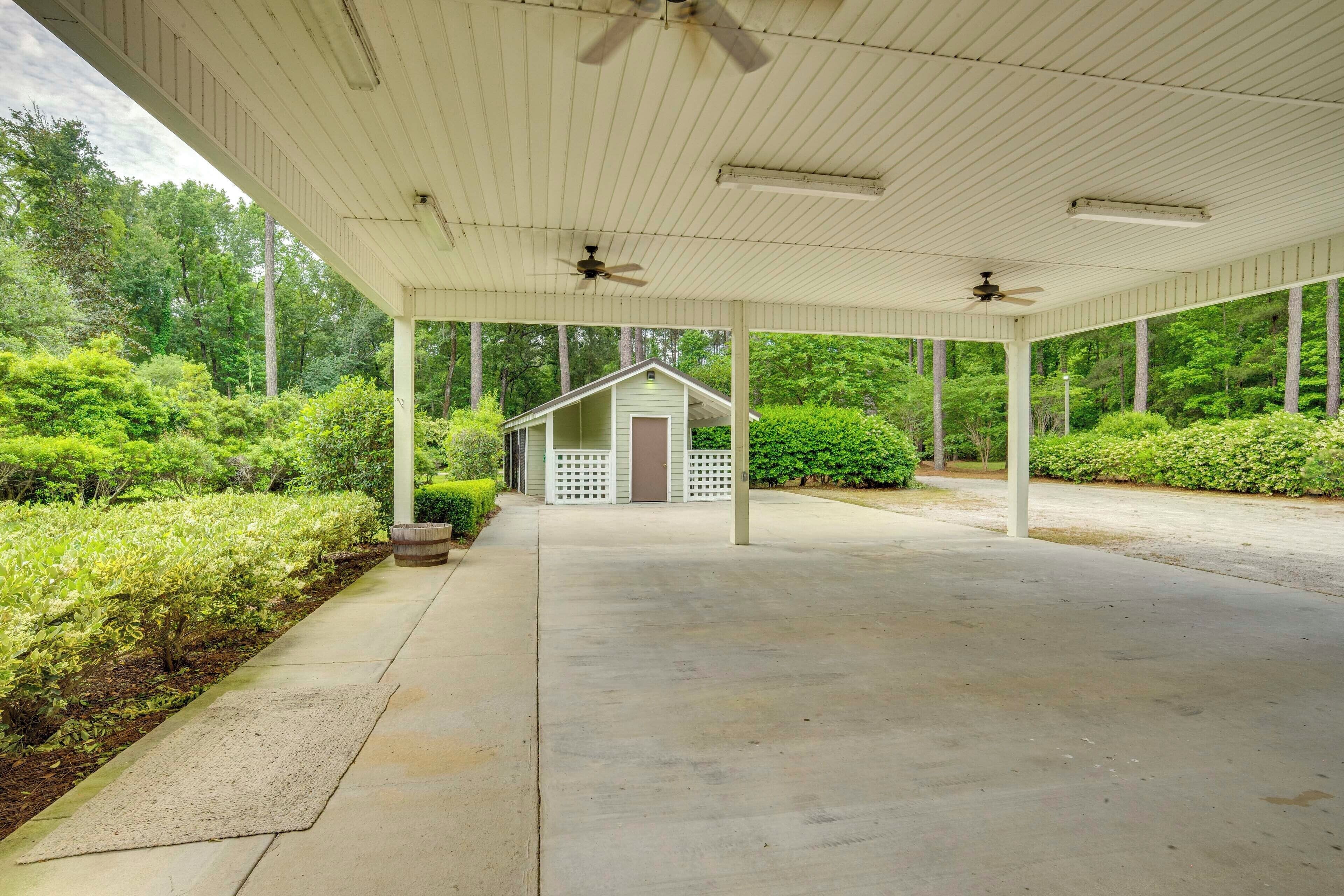 A covered outdoor area with a concrete floor, surrounded by greenery and a small building with a lattice front.