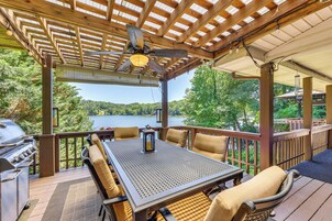 A covered outdoor deck with a table and chairs, a grill, and a view of a lake.