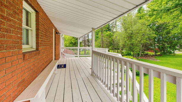 Une véranda couverte avec une balustrade blanche, un mur de briques et une vue sur une pelouse verdoyante et des arbres.