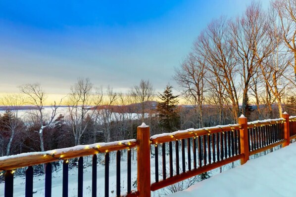 A wooden deck with snow-covered railings and string lights, overlooking a snowy landscape with trees and a distant view of a town.