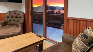 Ein Zimmer mit einer großen Schiebeglas-Tür, die auf einen Balkon mit Blick auf einen Sonnenuntergang über einer Berglandschaft führt.