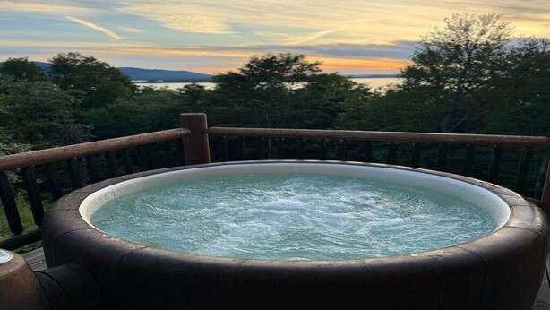 Ein Whirlpool auf einer Holzterrasse mit Blick auf Bäume und einem Sonnenuntergang im Hintergrund.