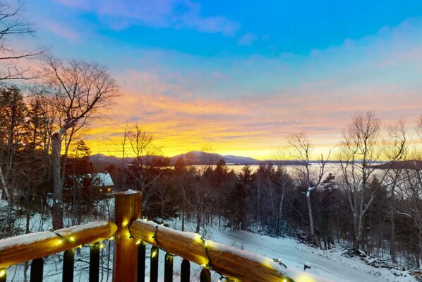 A snowy landscape with a wooden fence and string lights, overlooking a body of water and distant mountains.