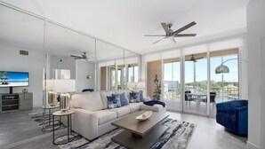 A modern living room with a sectional sofa, a coffee table, and a TV. There is a large window with a view of the city. - 🌴Great Water Views, Fabulous Location-Palm Harbor #403🌴 (Fort Myers Beach)