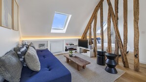A modern loft bedroom with a sloped ceiling, exposed wooden beams, a skylight, a blue sofa, a wooden coffee table, and a TV on a white stand. - Miel, 1BR/1BA, 3 people (París)